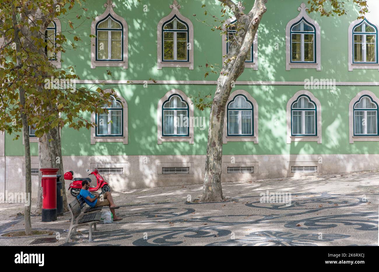 Uomo seduto con tablet su panca pubblica con scooter a motore, postbox rosso in piazza Bario Alto, Lisbona, Portogallo Foto Stock