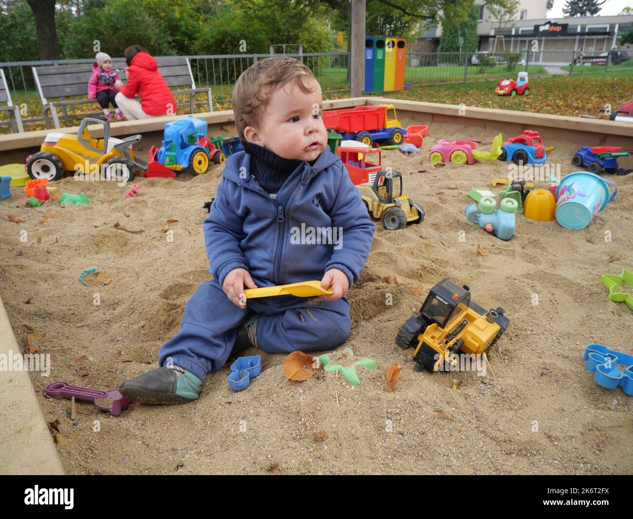 Un piccolo e carino bambino che gioca sulla sabbia in un parco giochi. Bambino seduto in un box di sabbia che tiene un giocattolo di plastica, con altri giocattoli sullo sfondo. Foto Stock
