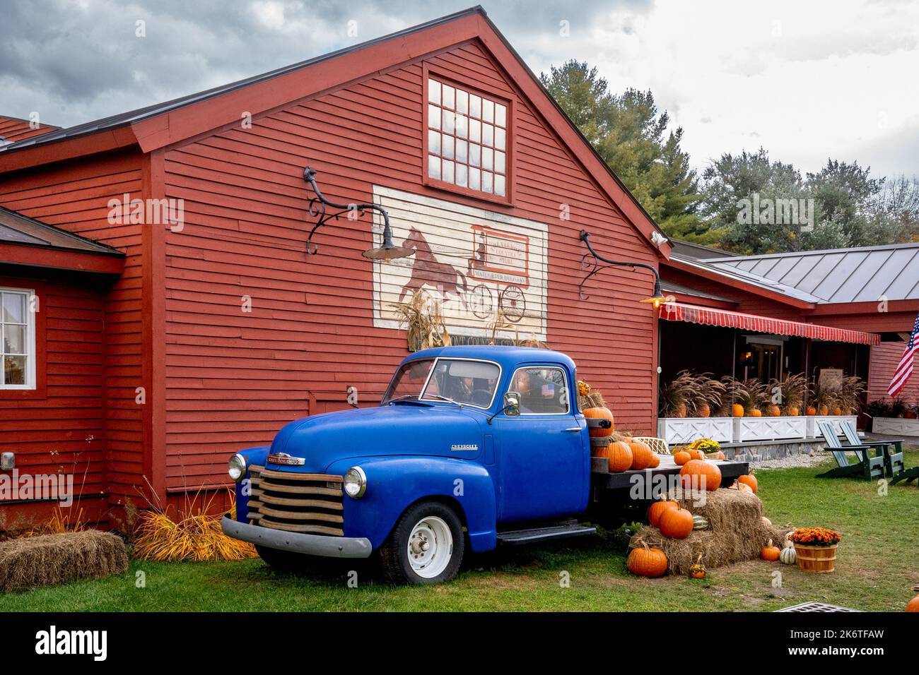 Weston, VT - USA - 7 ottobre 2022 esposizione orizzontale di zucche, fiori, zucche e paglia su un classico pickup americano blu parcheggiato lungo il bosco rosso Foto Stock