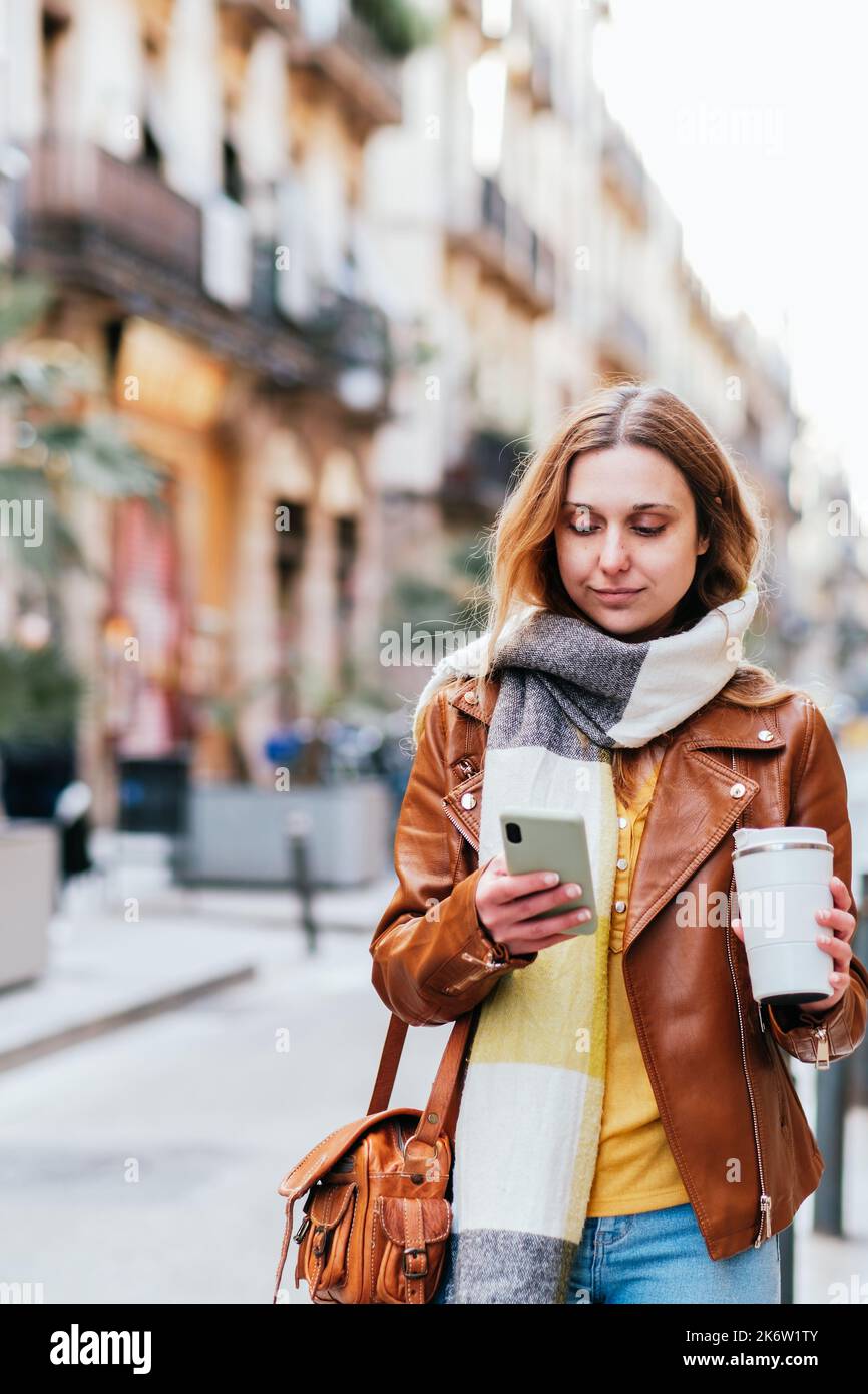 Donna focalizzata in giacca di pelle con caffè da asporto in mano messaggi di testo sul cellulare mentre si è in piedi sul sentiero in città Foto Stock