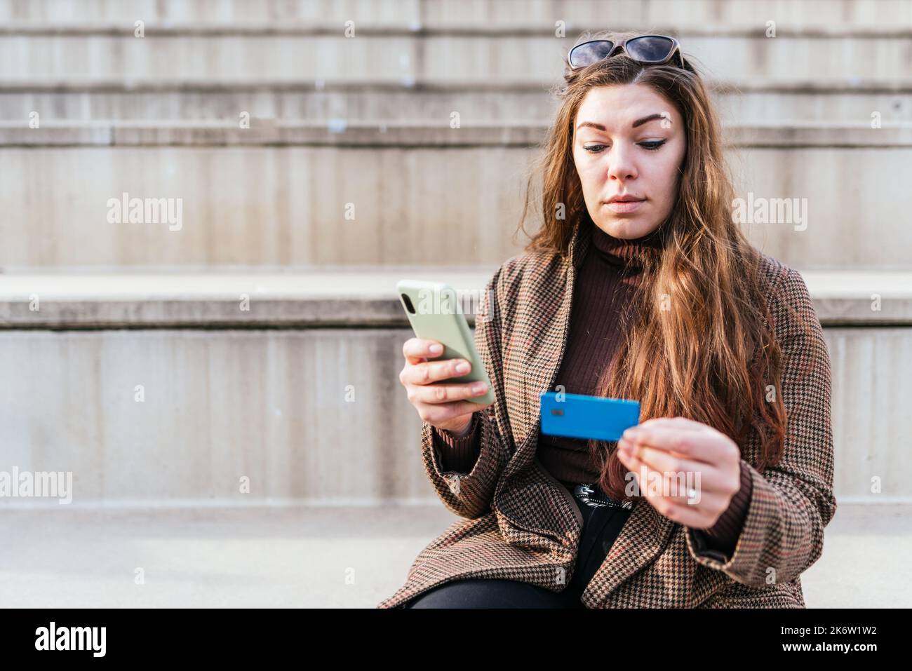 Donna in cappotto a scacchi che immette le credenziali della carta di credito nel cellulare mentre si siede sulle scale e facendo l'ordine in linea sulla strada della città Foto Stock