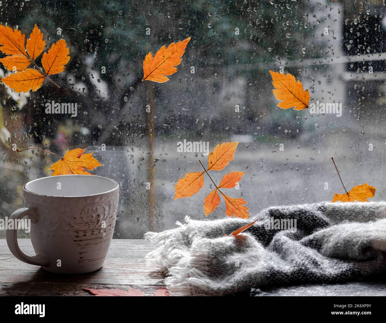 Gocce di pioggia e foglie cadute sulla finestra. Il tempo è tipico dell'autunno. Sfondo della finestra autunnale. Una tazza e un plaid o una sciarpa sulla finestra. Accogliente Foto Stock