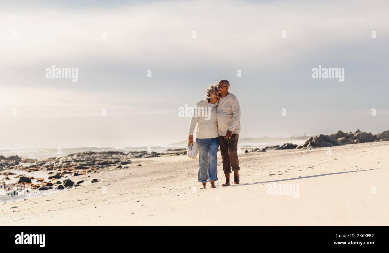 Romantica coppia anziana sorridente e abbracciante mentre si cammina a piedi nudi sulla spiaggia di sabbia. Coppia anziana felice godendo spendere un certo tempo di qualità Foto Stock