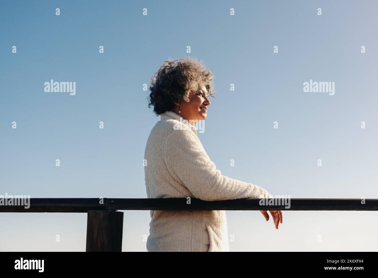 Donna matura che guarda una vista dell'oceano mentre si trova in piedi su una passerella in spiaggia. Felice donna anziana godendo una vacanza rinfrescante sul mare dopo Foto Stock
