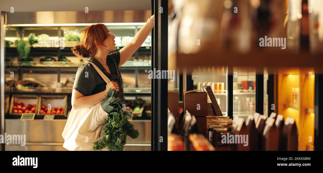 Cliente femminile che sceglie i prodotti alimentari da uno scaffale mentre trasporta una borsa con le verdure in un negozio di alimentari. Giovane donna che fa un po 'di shopping alimentari io Foto Stock
