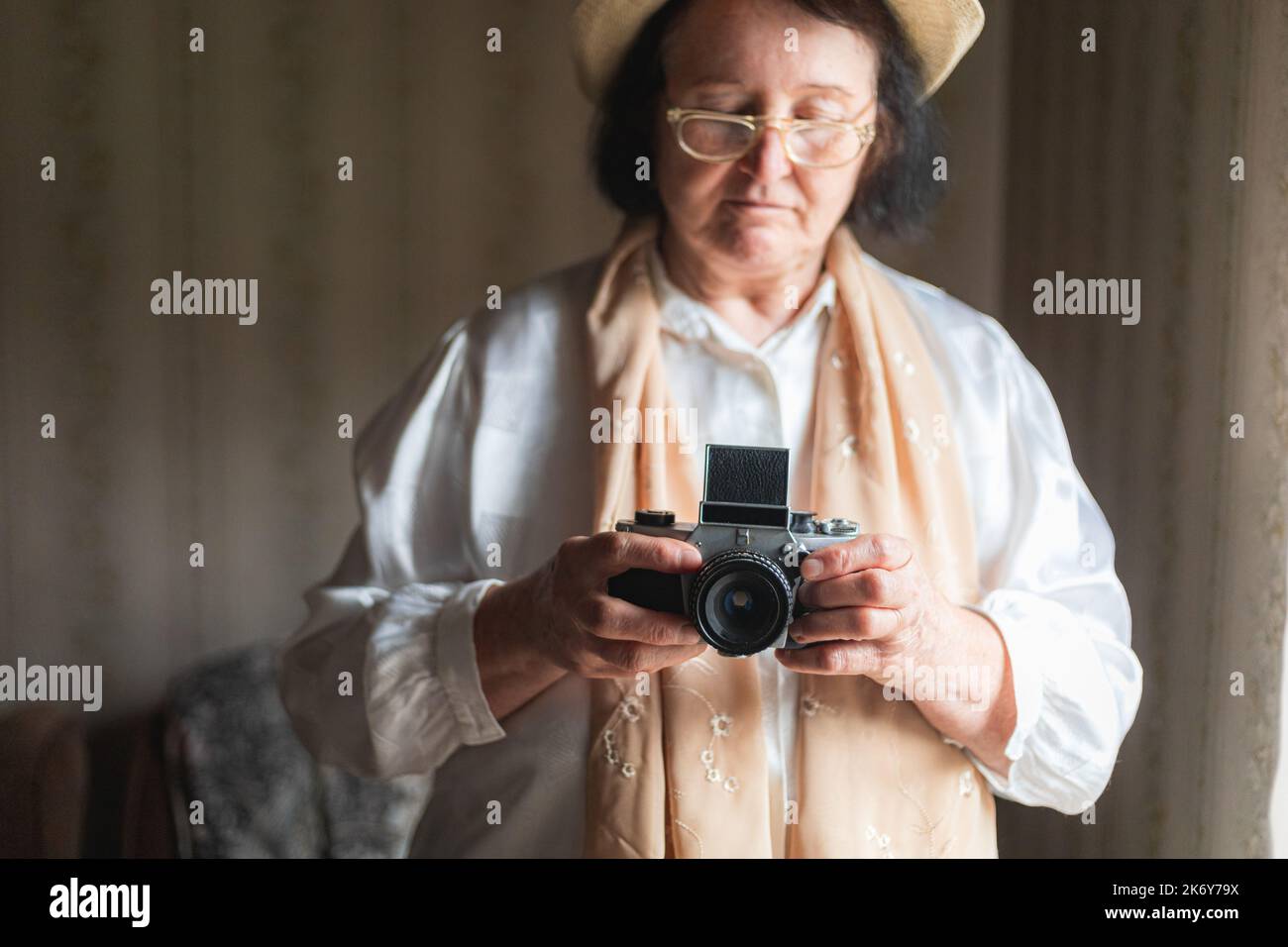 Donna anziana che tiene una macchina fotografica retrò, in piedi accanto alla finestra Foto Stock