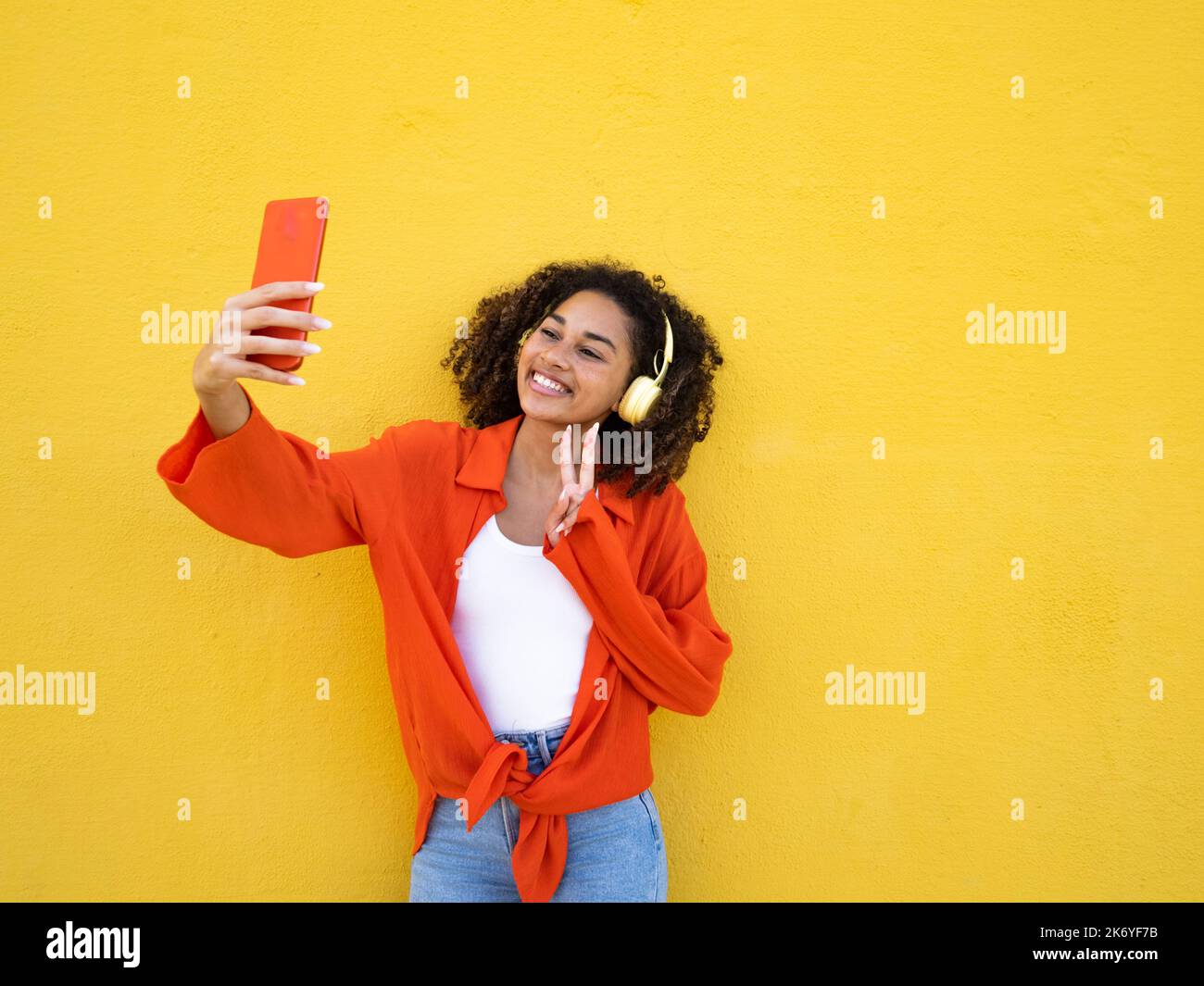 Allegra giovane donna afroamericana indossando cuffie che rendono simbolo di pace e prendendo un selfie sopra la parete gialla Foto Stock