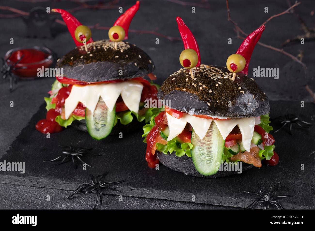 Cibo creativo di Halloween - due divertenti hamburger neri mostro con pesce, formaggio tostato, cetriolo, pomodoro, ketchup, olive occhi e corna peperoncino Foto Stock