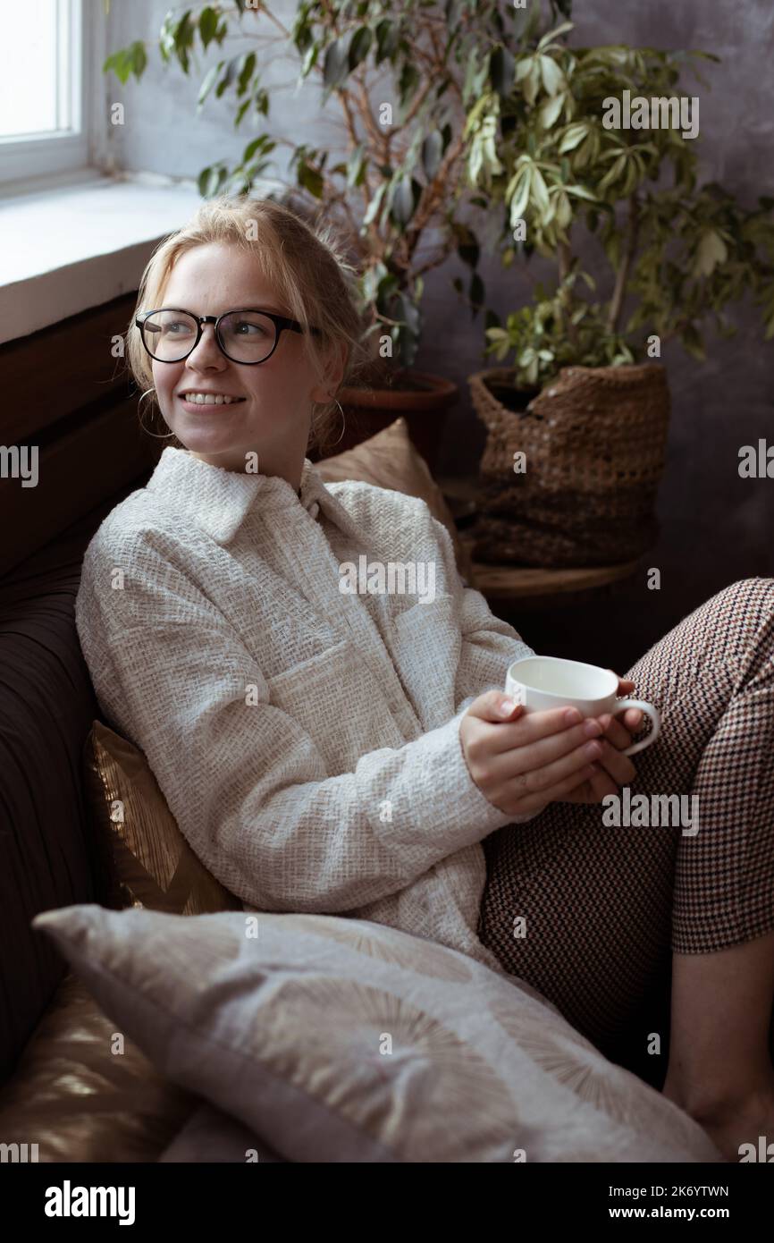 Donna bionda sorridente positiva verticale in bicchieri seduti su un comodo divano vicino a casa e tenere tazza, bere caffè o tè Foto Stock