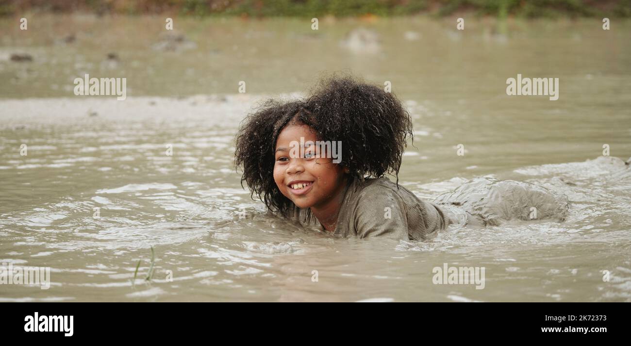 Ragazza felice afroamericana che gioca in pozza di fango bagnato durante piovendo nella stagione piovosa. Foto Stock