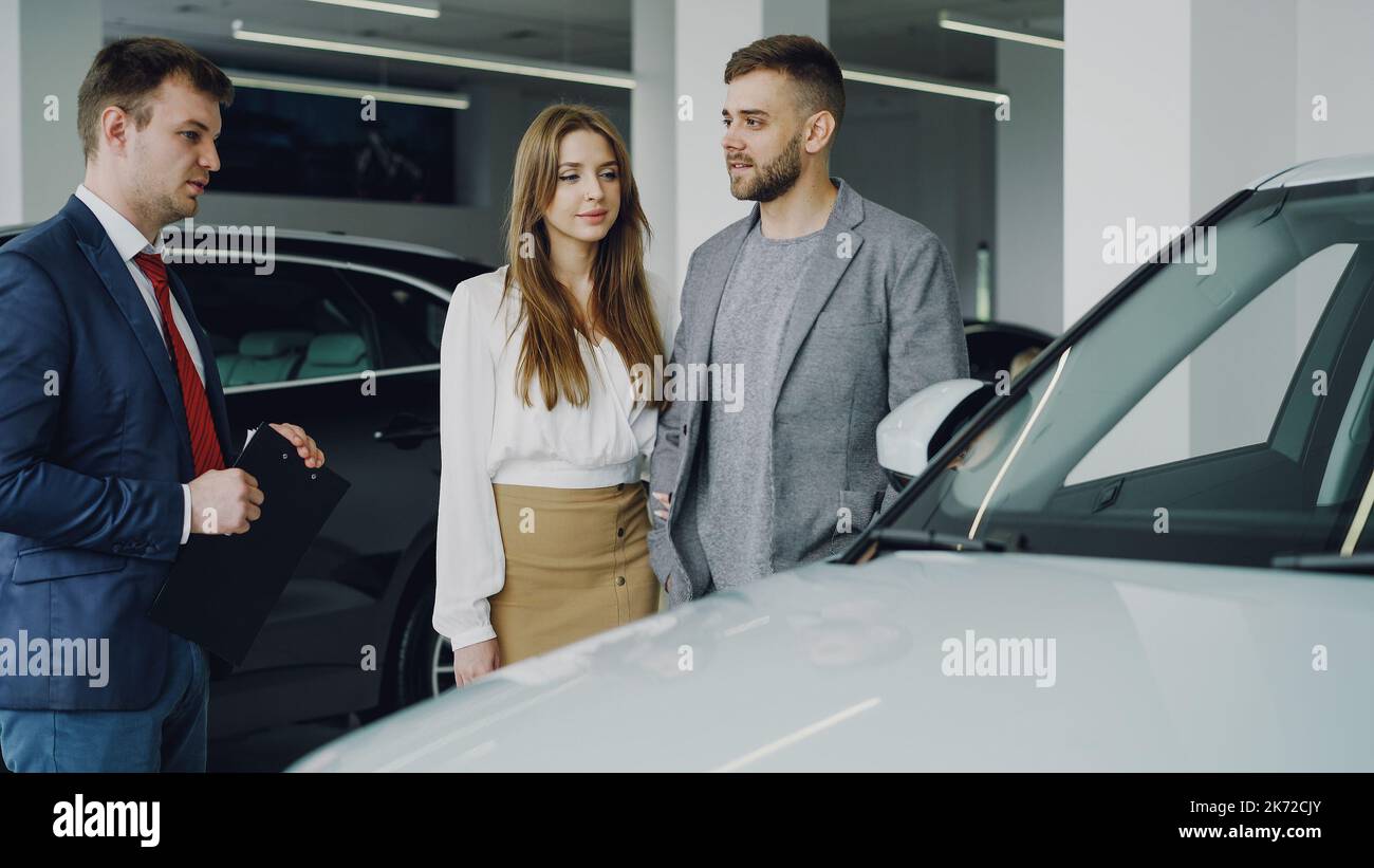 Il venditore amichevole dell'automobile in vestito sta comunicando con il giovane uomo sicuro che gli dice circa il nuovo modello del veicolo mentre la donna graziosa sta levandosi in piedi vicino al suo marito che tiene il suo braccio e sorridente. Foto Stock