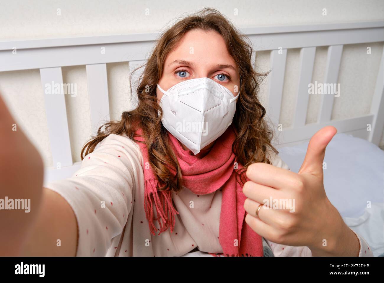 Donna malata blogger prende selfie foto sul telefono per il blog online. Donna ammalata adulta avvolta in una sciarpa rossa siede su un letto domestico in una camera da letto bianca, femmina A. Foto Stock