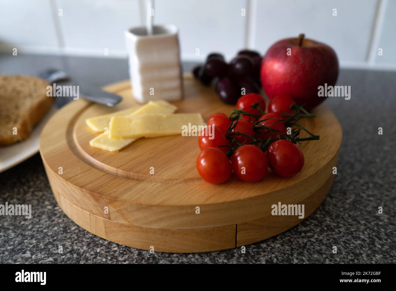 Un tagliere di formaggio con formaggio a fette, pomodori ciliegini, una mela rossa, mela, uva rossa e una pentola con cucchiaio, su un piano di lavoro della cucina con un piatto di raccolta Foto Stock