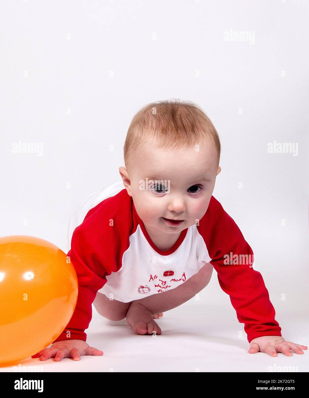Un bambino che guarda la macchina fotografica indossando un babygrow rosso e bianco che gioca con un palloncino arancione su uno sfondo bianco studio Foto Stock