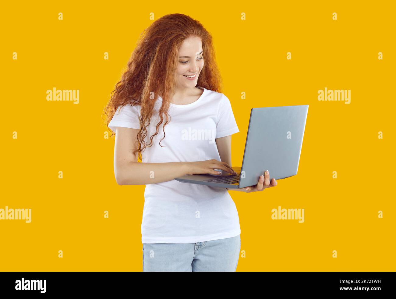 Ragazza sorridente arrossata con capelli ricci lunghi in abiti casual che lavorano su computer portatile su sfondo giallo. Foto Stock