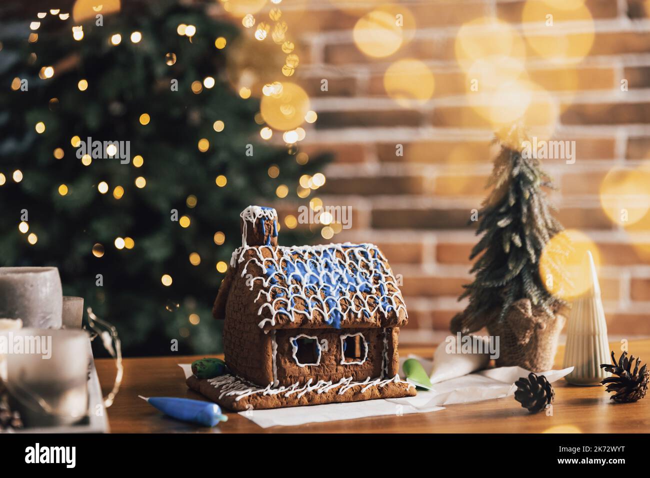 Piccola casa di pan di zenzero fatta in casa in piedi sul tavolo con decorazioni, candele nel soggiorno decorato con luci di Capodanno sfocate, abete di Natale. Meraviglioso umore felice di vacanza. Spazio di copia Foto Stock