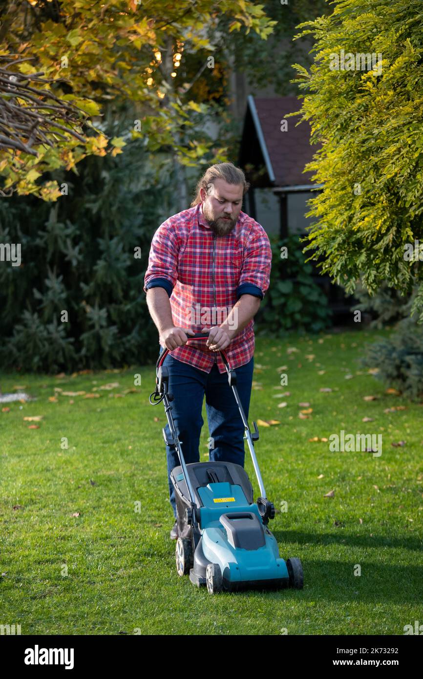 Bell'uomo con barba che spinge il rasaerba e rifilare l'erba nel suo cortile in autunno Foto Stock