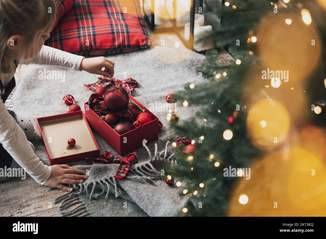 Bambina addobba l'abete di Natale con palline ornamentali rosse. Piccolo capretto che tiene il regalo in mano prima di appenderlo all'albero con la luce, garland. Scatola con palline appoggiate sul pavimento con decorazioni Foto Stock