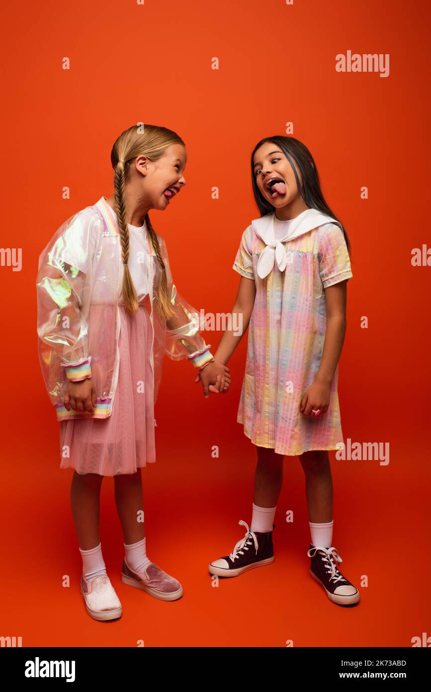 lunghezza completa delle ragazze divertenti che tengono le mani e che attaccano verso l'esterno le lingue a vicenda su fondo arancione Foto Stock