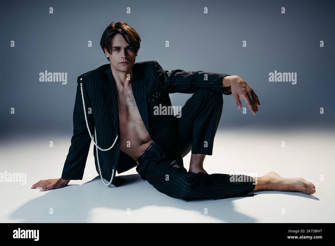 uomo a piedi nudi in abito elegante con perle e petto peloso seduto su sfondo grigio scuro Foto Stock