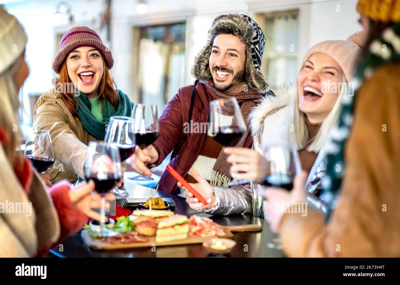 Giovani che brindano vino rosso al ristorante giardino - amici felici divertirsi insieme al bar della cantina che indossa abiti invernali Foto Stock