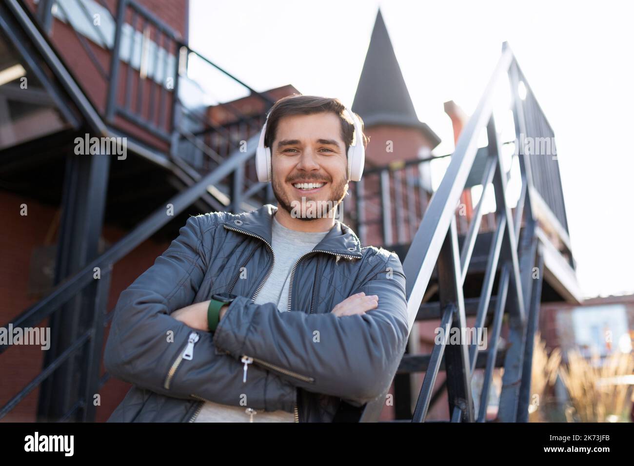 un uomo adulto con un ampio sorriso sul viso ascolta la musica in cuffia con le braccia incrociate sul petto sullo sfondo delle scale fuori Foto Stock