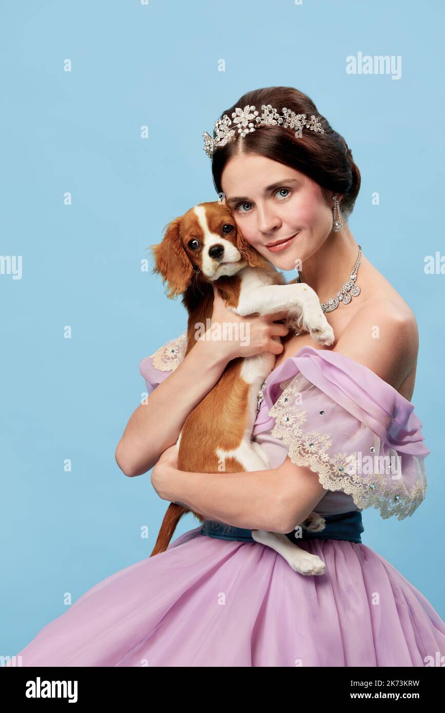 Amato animale domestico. Ritratto di giovane donna affascinante in lilla colore vintage vestito come principessa medievale tenendo piccolo re Carlo spaniel cane sul blu Foto Stock