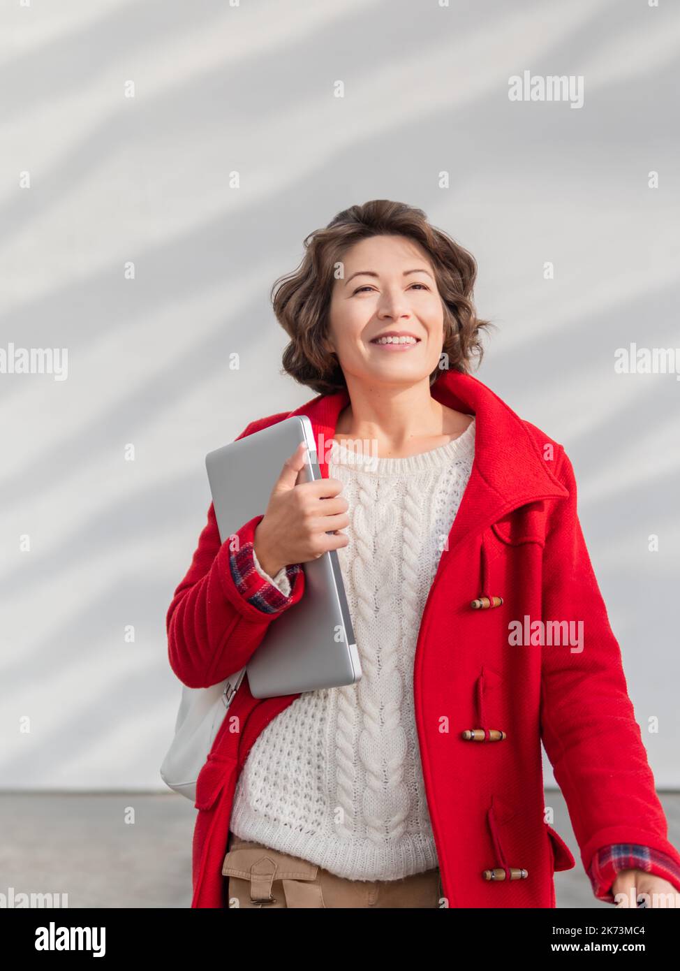 Donna ricciola in mantello rosso è in piedi da parete bianca. Studente adulto sorridente con un computer portatile. Freelance con dispositivo portatile per il lavoro. Stile di vita moderno. Foto Stock