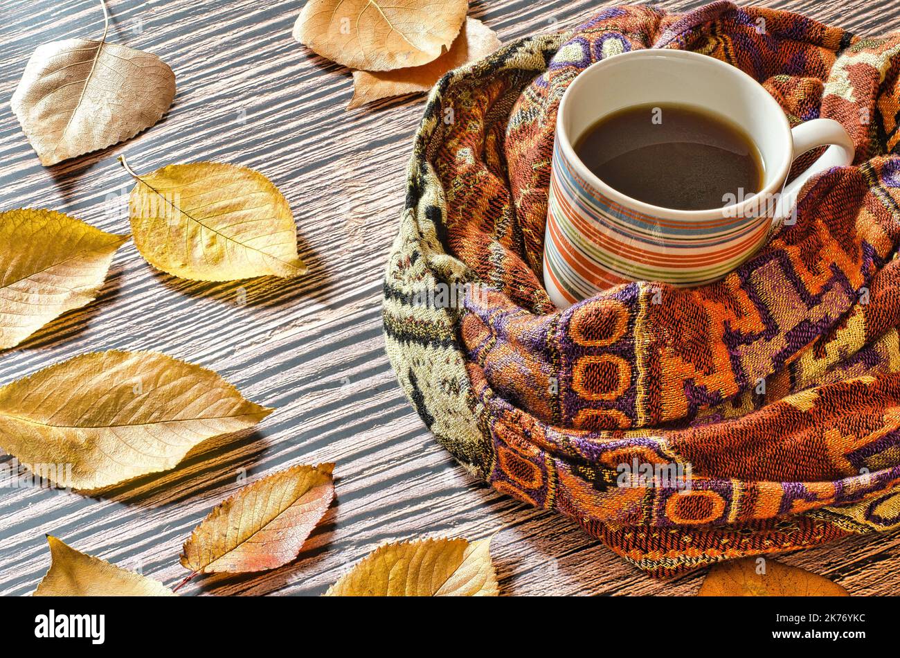 tazza di caffè caldo avvolto in una sciarpa su un tavolo di legno con foglie autunnali cadute Foto Stock