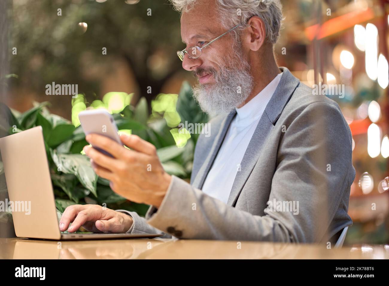 Felice uomo d'affari anziano che tiene in mano uno smartphone utilizzando un notebook che lavora tardi. Foto Stock