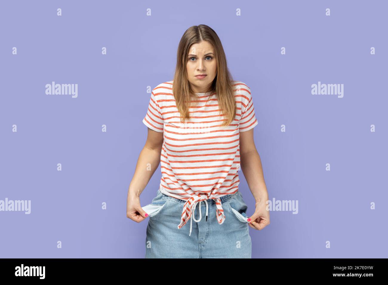 Disoccupazione e fallimento. Ritratto di donna bionda che indossa T-shirt a righe che si sfogli tasche vuote, preoccupato per i debiti, nessun denaro per vivere. Studio al coperto isolato su sfondo viola. Foto Stock