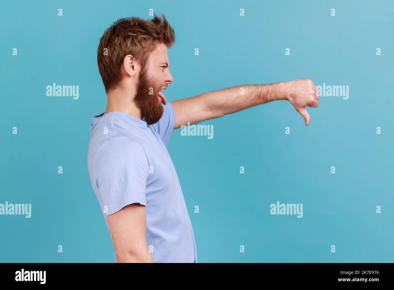 Vista laterale dell'uomo bearded che critica la qualità difettosa con i pollici giù la grimace dispiaciuta, che mostra il gesto dislike, che esprime la disapprovazione. Studio in interni isolato su sfondo blu. Foto Stock