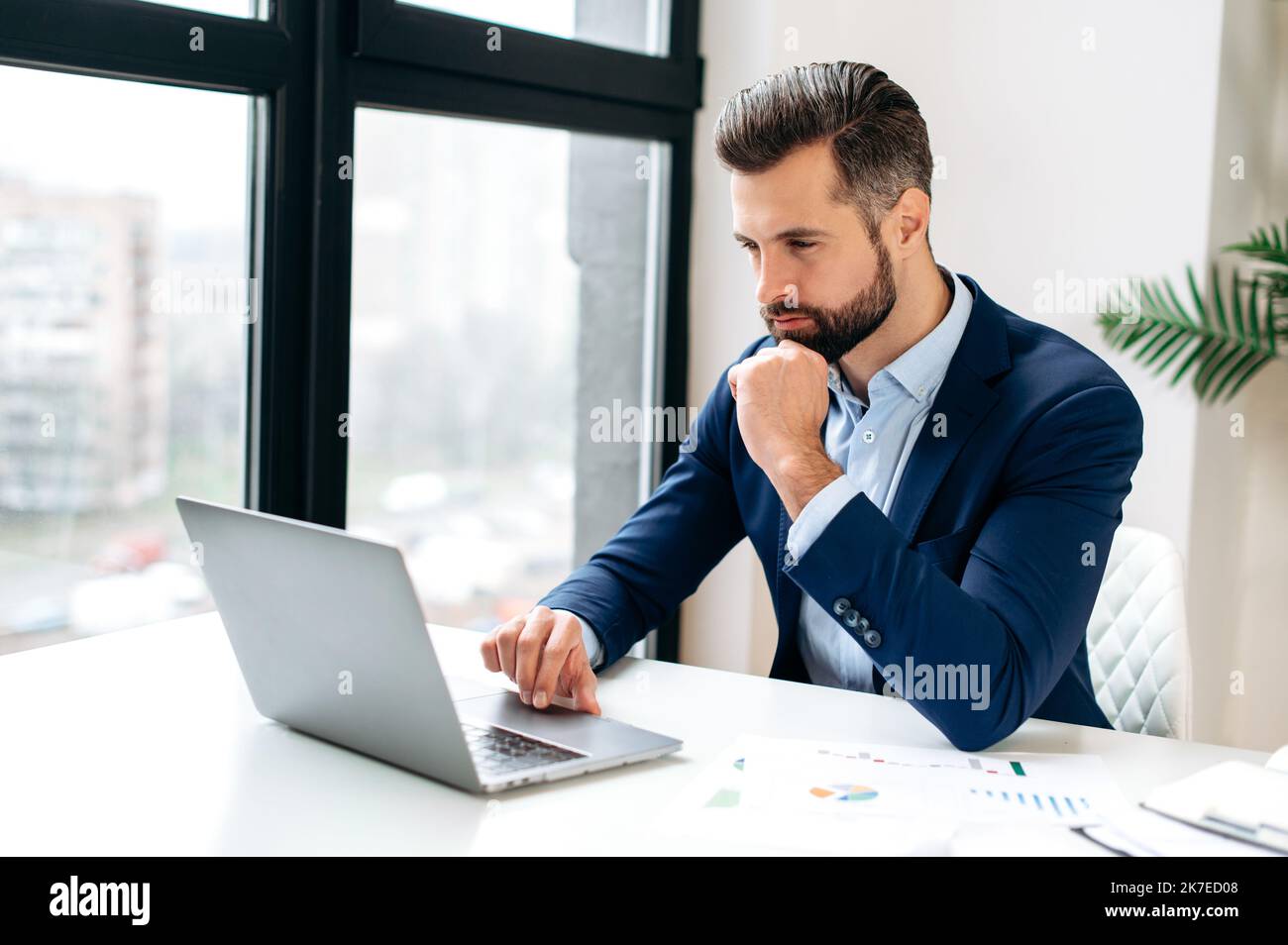 Focalizzato riuscito sicuro occupato uomo d'affari caucasico in un vestito formale, si siede sul posto di lavoro, lavorando su un progetto utilizza laptop, scrivendo strategia di business, analizzando le informazioni, navigando in Internet Foto Stock