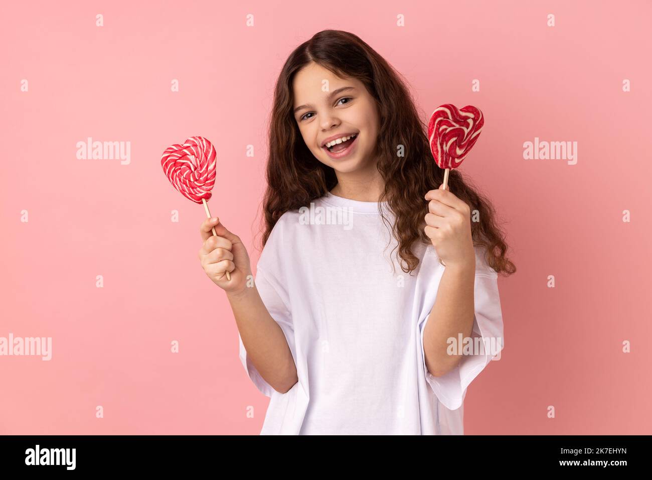 Ritratto di piccola ragazza soddisfatta che indossa una T-shirt bianca con due lollypops a forma di cuore, guardando la fotocamera con felice espressione. Studio in interni isolato su sfondo rosa. Foto Stock