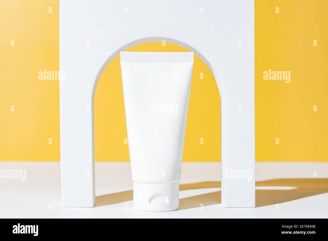 Tubo bianco per cosmetici in plastica e podio bianco ad arco su sfondo giallo. Cura della pelle, trattamento di bellezza, concetto spa. Vetrina alla moda di cosmetici p Foto Stock