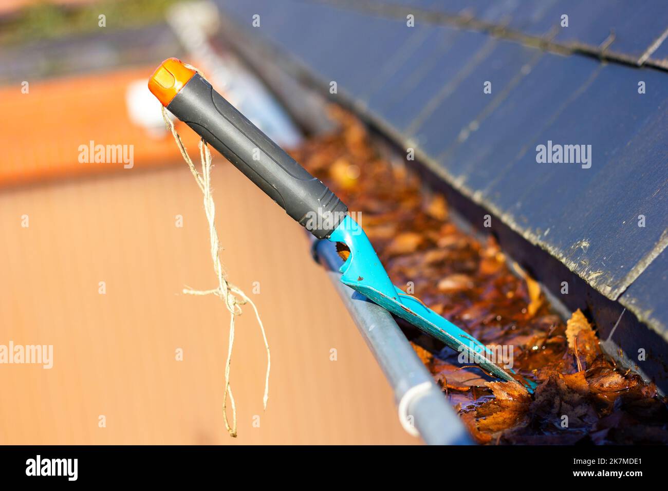 Un ritratto di una grondaia del tetto piena di foglie autunnali colorate cadute in una giornata di sole con una piccola pala da giardino blu in esso di qualcuno che sta pulendo il Foto Stock