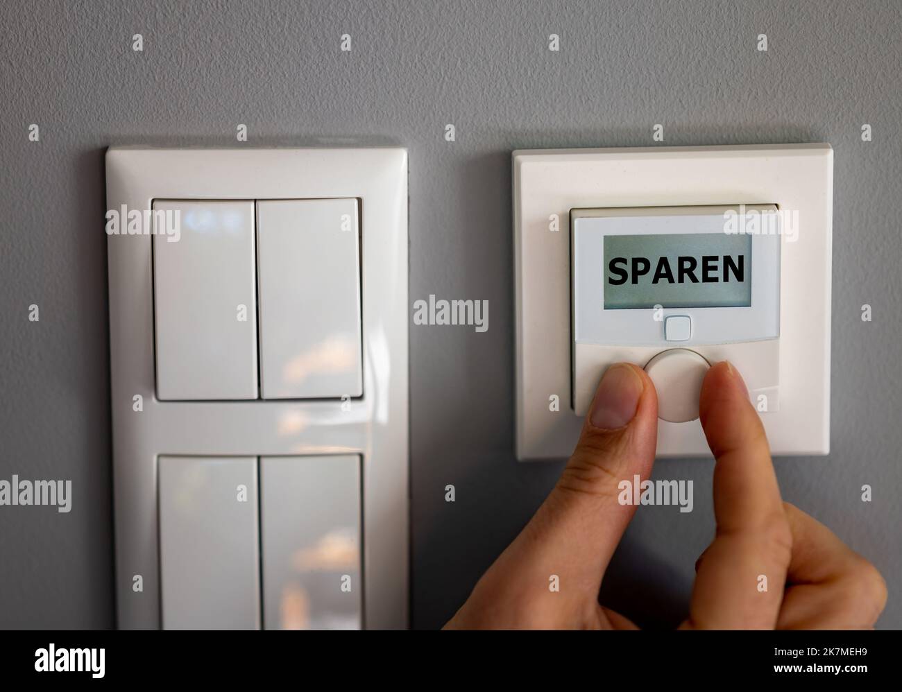 La mano modifica la temperatura su un termostato elettronico. Sul display viene visualizzata la parola tedesca 'paren' (salvataggio). Simbolo per il risparmio energetico. Foto Stock