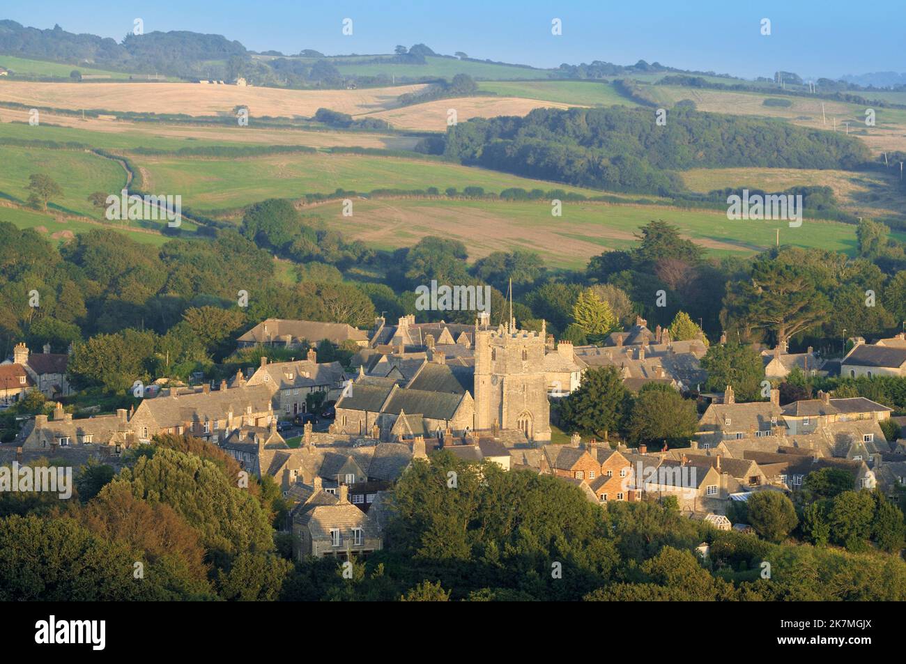 Vista panoramica della torre della chiesa di St Edwards, la chiesa parrocchiale nel villaggio del castello di Corfe, con le case che si trovano contro la campagna nella tarda estate, Dorset, Inghilterra, Regno Unito Foto Stock
