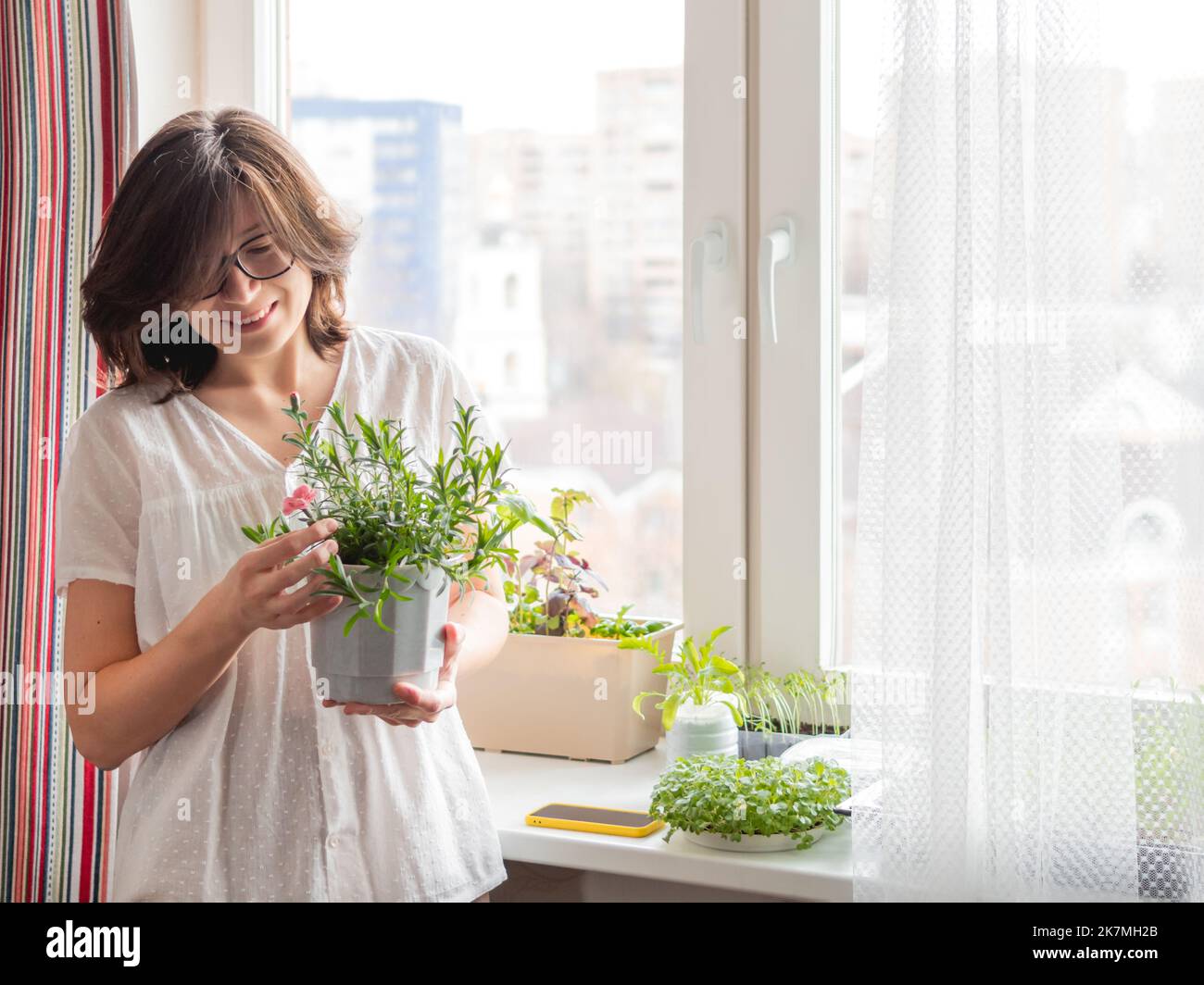 Donna sorridente sta guardando sul fiore di garofano fiorente.. Coltivazione di basilico organico commestibile, rucola, microgredo di cavolo per una sana alimentazione. Giardinaggio Foto Stock