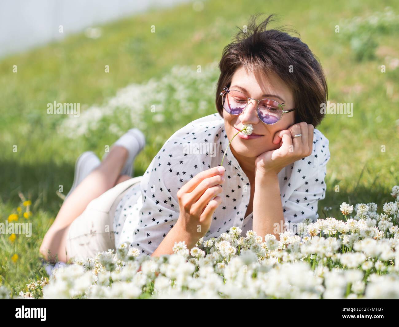 Donna in occhiali da sole colorati sniffs fiori trifoglio sul prato in parco urbano. Natura in città. Rilassati all'aperto dopo il lavoro. Atmosfera estiva. Foto Stock