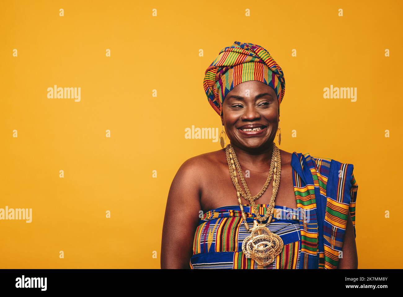 Donna ghanaiana sorridente che indossa un abito tradizionale su sfondo giallo. Felice donna nera vestita in tessuto colorato Kente e gioielli d'oro Foto Stock