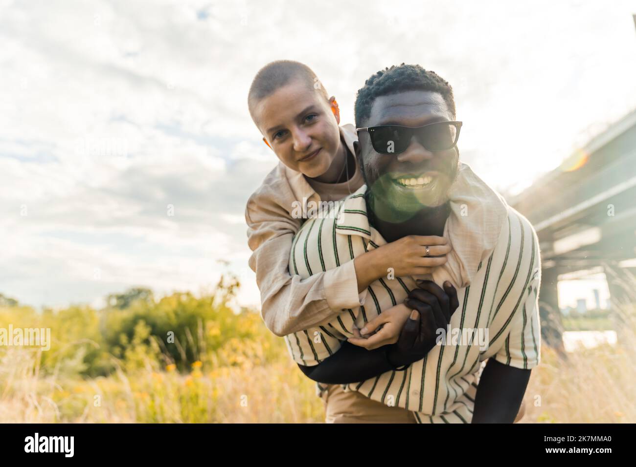 Splendido ritratto all'aperto di coppie multietniche che si godono il tempo all'aperto. Bell'alto uomo nero in occhiali da sole sorridente alla macchina fotografica, prendendo la sua ragazza calva caucasica sul piggyback. Foto di alta qualità Foto Stock