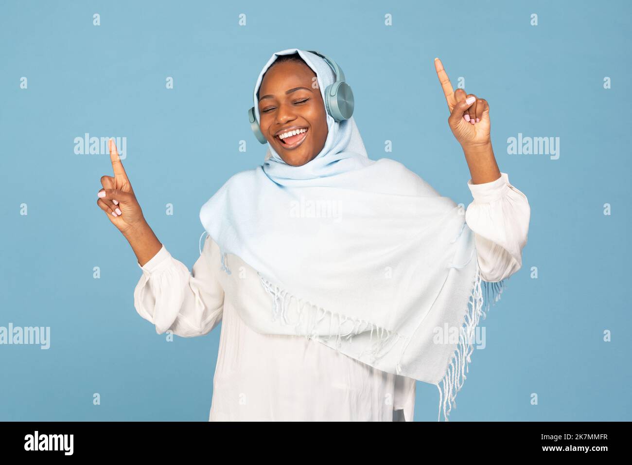 Donna musulmana afro-americana overjoyed in hijab divertirsi, ballare e cantare con mani sollevate, sfondo blu Foto Stock