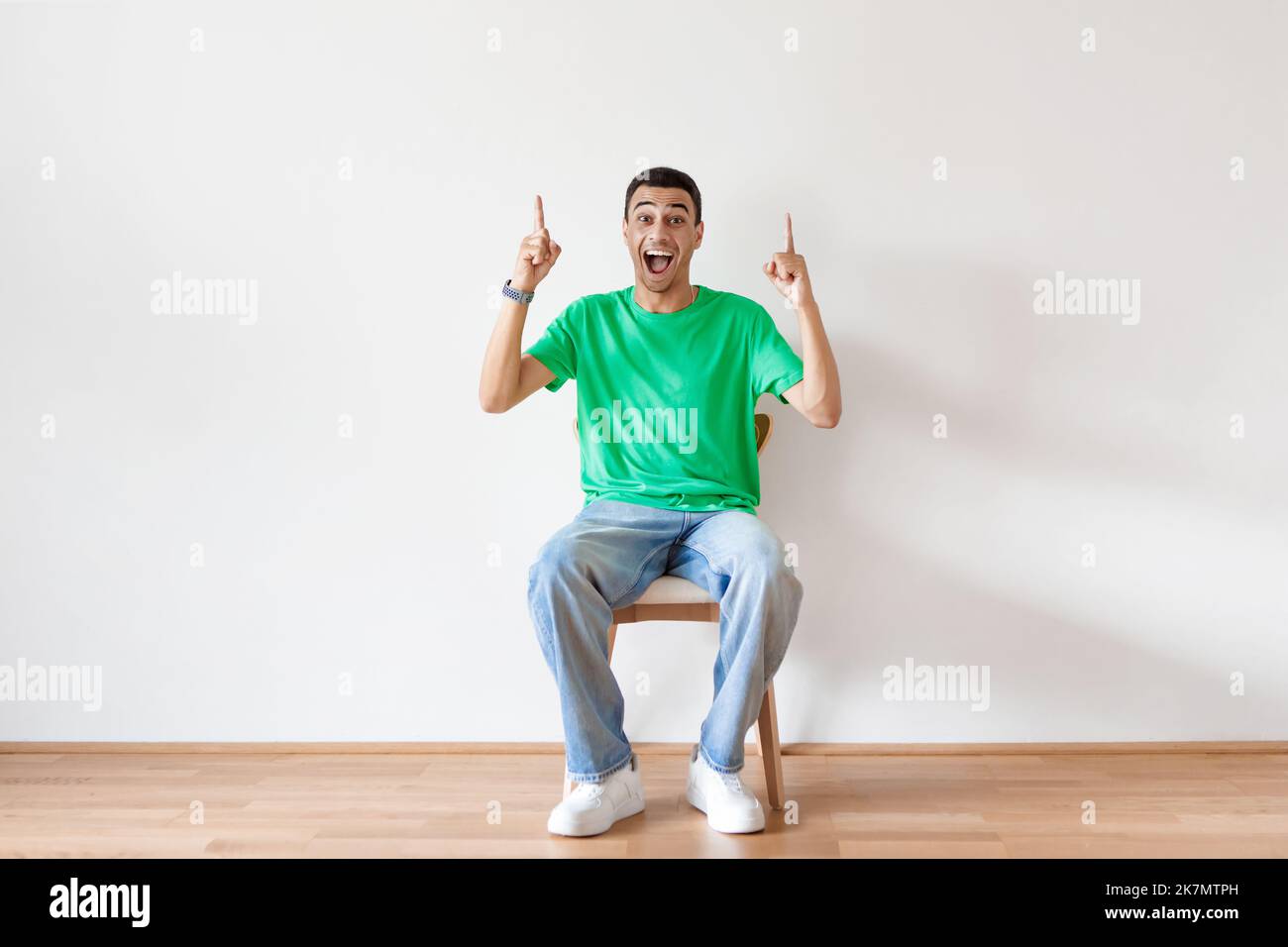 Uomo arabo overjoyed seduto sulla sedia e puntando due dita in su, offrendo spazio per la vostra pubblicità, parete di luce Foto Stock