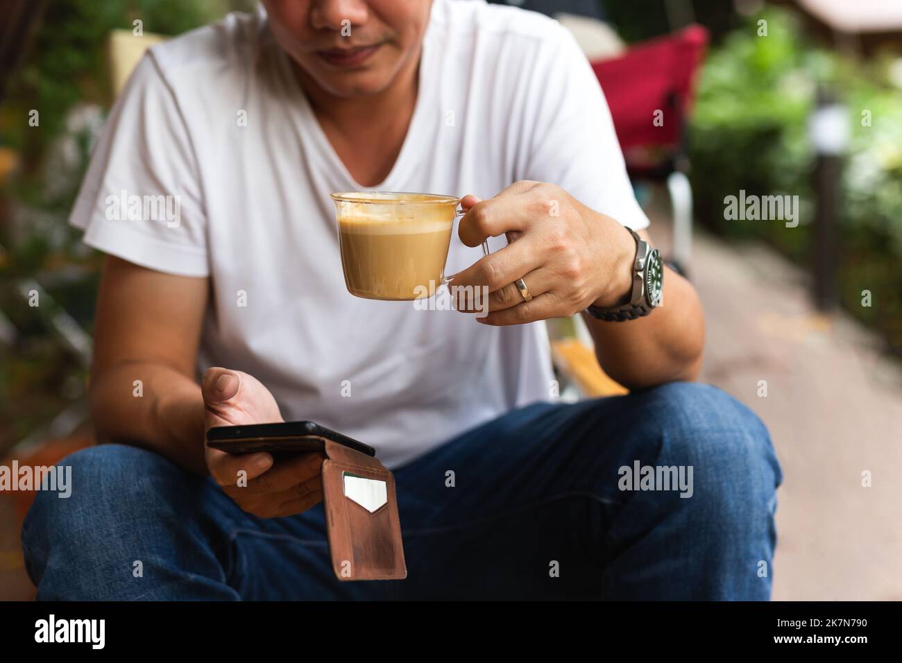 Uomo in jean e t shirt bianca bere caffè e guardare il telefono cellulare in caffè all'aperto. Foto Stock