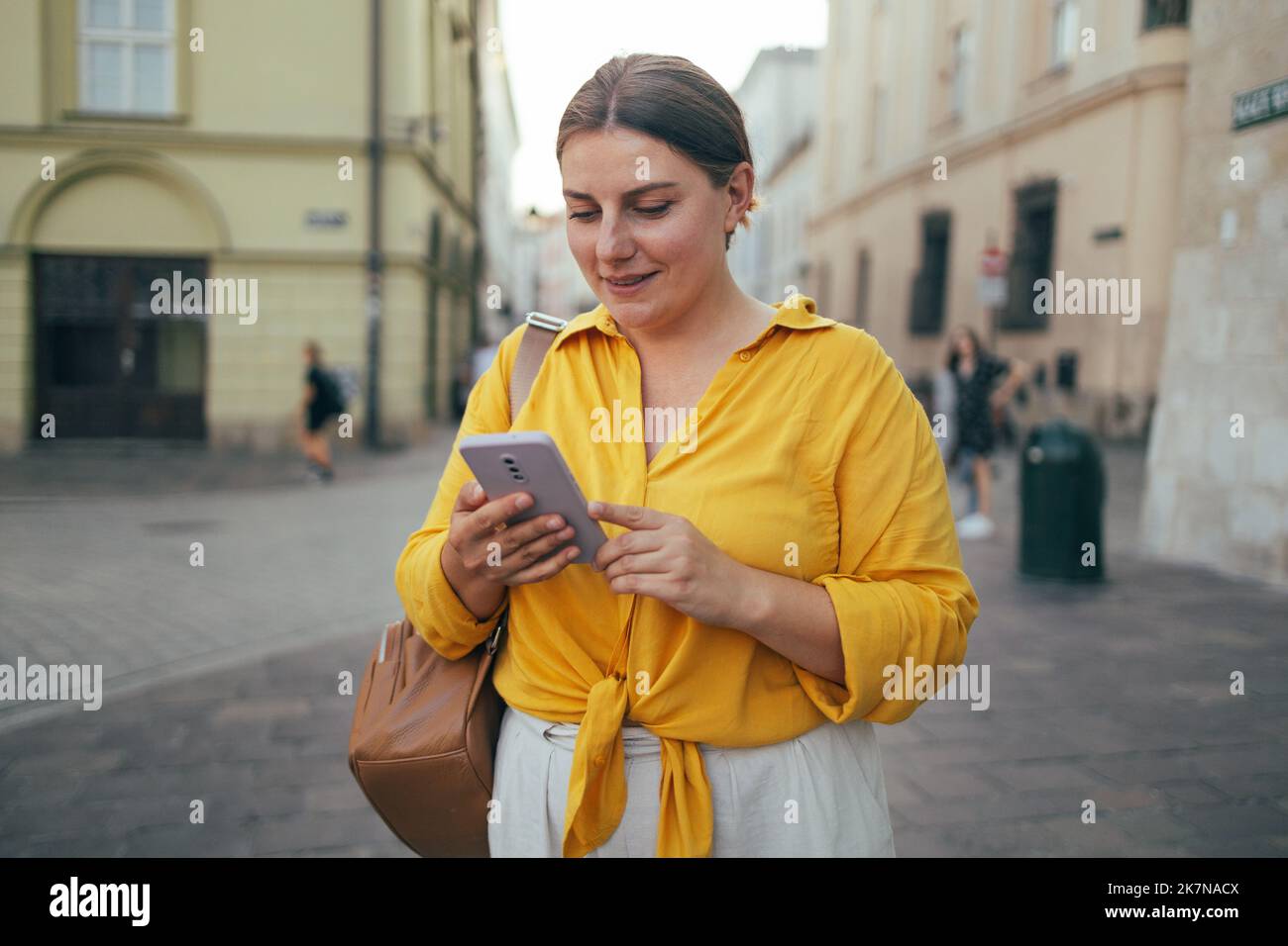 Donna che indossa una maglietta gialla arancione con un SMS sullo smartphone che cammina per strada in una giornata di sole Foto Stock