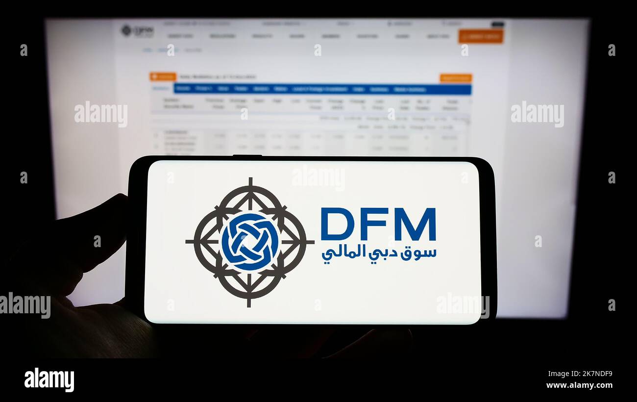 Persona che tiene il cellulare con il logo della borsa valori Dubai Financial Market (DFM) sullo schermo di fronte al sito web aziendale. Messa a fuoco sul display del telefono. Foto Stock