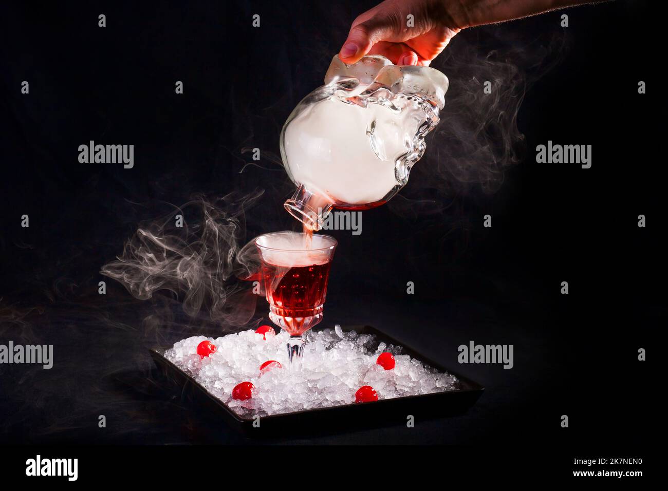Il barista versa un cocktail alcolico affumicato da una bottiglia a forma di cranio, che l'autore serve su un piatto con ghiaccio tritato e ciliegie. Foto Stock