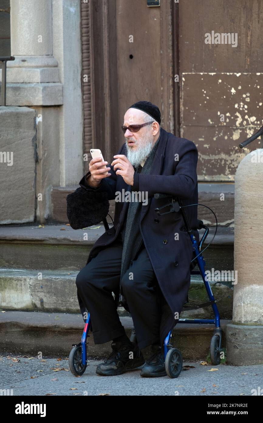 Un più anziano ebreo si siede sul suo camminatore e a sembra essere un raro uomo hassidico che usa uno smartphone. A Brooklyn, New York. Foto Stock