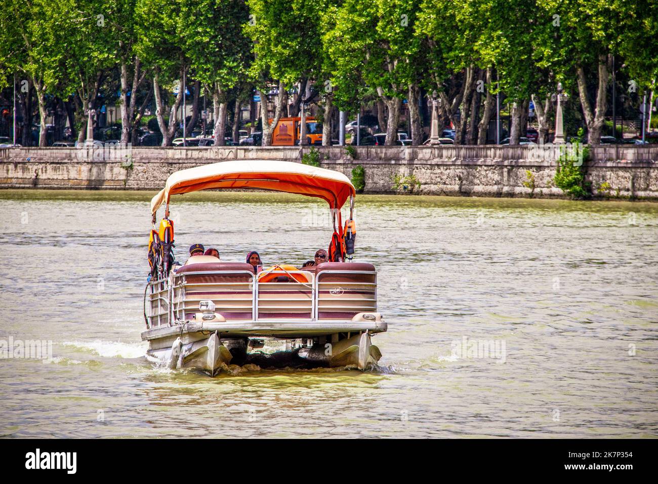 2019 07 2019 Tbilisi Georgia - Pontoon barca con passeggeri femminili in velo su Mtkvari - fiume Kura con muro di contenimento e alberi sullo sfondo Foto Stock